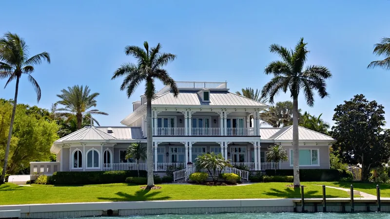 Light gray two story Florida home with white porches, a metal roof and tall palm trees above a green lawn